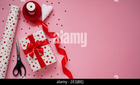Geschenkverpackung zum Valentinstag. Box in weißem Papier in Herzen, rote Schleife, Süßigkeiten, rotes Band, Schnitter und eine Rolle Geschenkpapier auf einem rosa gewickelt Stockfoto