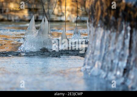 Nahaufnahme von Eiszapfen, die an Wurzeln, Ästen und Felsen hängen, die sich neben einem Bach bildeten. Stockfoto