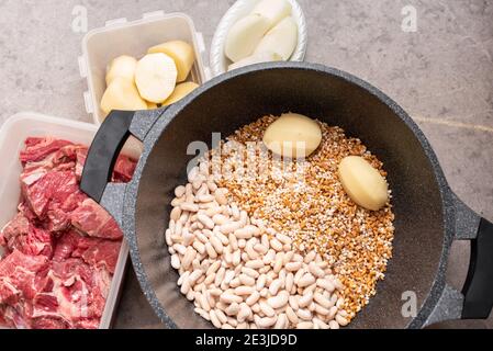 Nahaufnahme des traditionellen jüdischen Cholent Hamin - Hauptgericht für die Schabbat Mahlzeit langsam gekochtes Rindfleisch mit Kartoffeln, Bohnen, Weizenkörnern und Gewürzen in einer Kasserolle auf einem grauen Marmor Küchentisch. Stockfoto