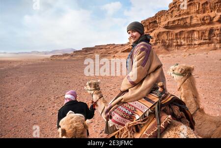 Junge Frau, die in der Wüste Wadi Rum auf einem Kamel reitet, lächelnd über ihre Schulter blickt. Es ist ziemlich kalt, so trägt sie traditionellen Beduinenmantel - bis Stockfoto