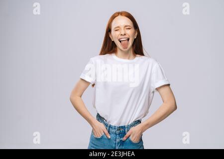 Fröhliche junge Frau trägt T-Shirt und Denim-Hosen zeigen Zunge auf isolierten weißen Hintergrund. Stockfoto