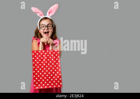 Frohe Ostern. Ein fröhliches kleines Mädchen in einem rosa Kleid mit Tupfen hält eine Einkaufstasche in den Händen. Nettes Baby auf einem grauen Hintergrund. Stockfoto