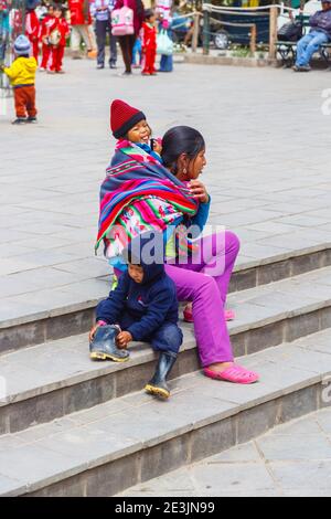 Eine einheimische Frau, die ihr Baby und ein Kind trägt, sitzt auf einer Treppe auf dem Stadtplatz von Ollantaytambo im Heiligen Tal in Urubamba, Cusco Region, Peru Stockfoto