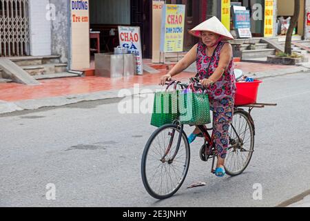 Vietnamesische Frau, die auf dem Fahrrad reitet, trägt traditionelle nón lá, asiatische konische Hut, in der Stadt Ninh Binh im roten Flussdelta von Nordvietnam Stockfoto