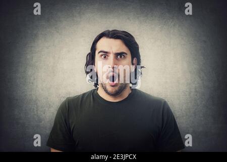 Porträt von schockierten jungen Mann, lange lockige Frisur, starrte mit großen Augen und Mund offen isoliert auf grauen Wand Hintergrund. Erstaunt Kerl, erstaunt ex Stockfoto