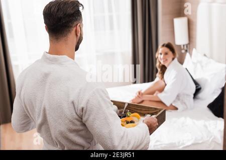 Mann im Bademantel hält Frühstückstablett neben Freundin im Hotel Bett auf unscharfem Hintergrund Stockfoto