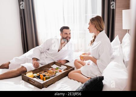 Lächelnder Mann im Bademantel, der die Freundin beim Frühstück ansah Tablett auf Hotelbett Stockfoto