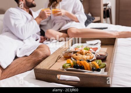 Kurzer Blick auf frisches Obst auf Frühstückstablett in der Nähe von Pärchen In Bademänteln auf verschwommenem Hintergrund auf Hotelbett Stockfoto