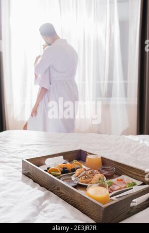 Frühstück auf Holztablett auf dem Bett und Paar in Bademänteln Umarmt nahe Fenster auf verschwommenem Hintergrund im Hotelzimmer Stockfoto