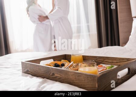 Kurzer Blick auf das köstliche Frühstück auf dem Tablett auf dem Bett und Paar in Bademänteln auf verschwommenem Hintergrund im Hotel Stockfoto