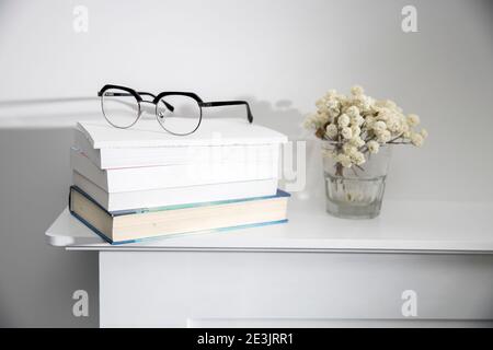Ein Stapel Bücher, Gläser darauf, auf einer weißen Tischplatte. Getrocknete Gipsophila Blumen in einer Glasvase in der Nähe. Speicherplatz kopieren Stockfoto