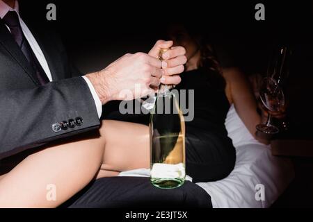 Mann öffnet Flasche Champagner in der Nähe Freundin auf dem Bett Unscharfer Hintergrund im Hotel während der Nacht Stockfoto