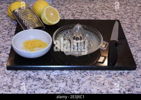 Zitronenpresse aus Glas und Zitronenreibe mit Zitronen und Zitronenschale in einer kleinen Schüssel auf einem schwarzen Granitbrett. Ordentliches Kochen und Speisenzubereitung. Stockfoto