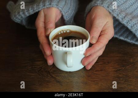 Die Hände der Frau im Pullover halten eine Tasse starken Kaffee auf dem Holztisch. Kaffeeventilator Draufsicht Hintergrund Stockfoto