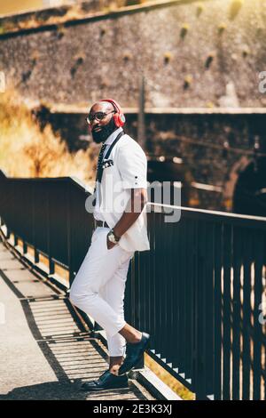 Ein ausgefallener reifer kahler schwarzer Mann in einem weißen eleganten Anzug und mit einem gepflegten Bart lehnt sich an ein Zaun Sie im Freien, während Sie Musik hören Stockfoto