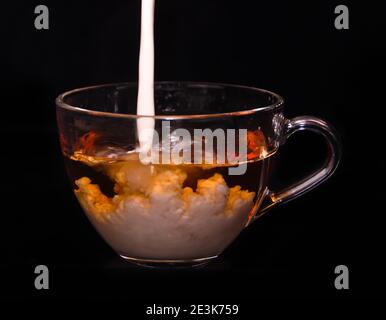 Weiße Milchcreme gießt in eine Tasse Tee und macht vor schwarzem Hintergrund einige Tropfen, Blasen und Wolken Stockfoto