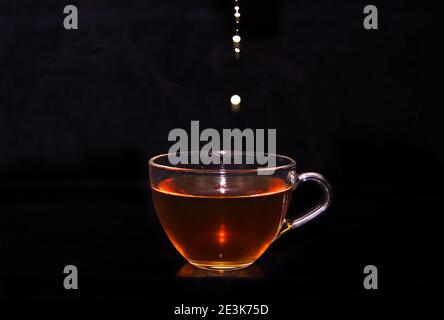 Weiße Milchcreme gießt in eine Tasse Tee und macht vor schwarzem Hintergrund einige Tropfen, Blasen und Wolken Stockfoto