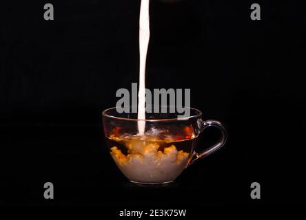 Weiße Milchcreme gießt in eine Tasse Tee und macht vor schwarzem Hintergrund einige Tropfen, Blasen und Wolken Stockfoto