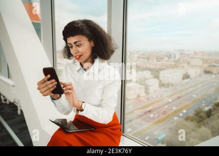 Eine schillernde junge afroamerikanische Geschäftsfrau mit einem digitalen Tablet Ihr roter Rock beantwortet eine Nachricht auf ihrem Smartphone Beim Sitzen in der Nähe von Th Stockfoto