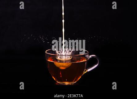 Weiße Milchcreme gießt in eine Tasse Tee und macht vor schwarzem Hintergrund einige Tropfen, Blasen und Wolken Stockfoto