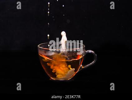 Weiße Milchcreme gießt in eine Tasse Tee und macht vor schwarzem Hintergrund einige Tropfen, Blasen und Wolken Stockfoto