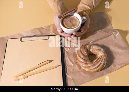 Hände Halten Tasse Tee oder Glühwein nahe Resume Blatt und Kuchen auf dem Set Sail Champagner Farbe Hintergrund, Draufsicht. Stockfoto