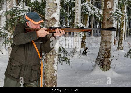 Im verschneiten Winterwald lehnt sich ein erfahrener, alter Jäger an einen Baum und hält sein Gewehr in der Schussposition. Stockfoto