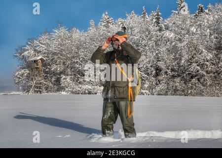 An einem schönen Wintermorgen steht ein Jäger im tiefen Schnee vor seiner hohen Jagdkanzel und beobachtet sein Jagdgebiet mit seinem Fernglas. Stockfoto