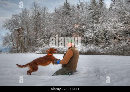 Ein Jäger spielt glücklich mit seinem jungen irischen Setter-Jagdhund im Schnee vor seiner Jagdkanzel. Stockfoto