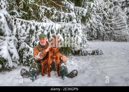 Im verschneiten Schwarzwald sitzt ein Jäger mit seinem schönen irischen Setter-Jagdhund am Waldrand im Schnee. Stockfoto