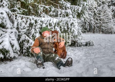 Enge Freunde. Im verschneiten Winterwald sitzt ein älterer Jäger mit seinem jungen irischen Setter-Jagdhund im schnee und wird zärtlich in den nos gebissen Stockfoto