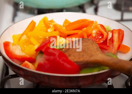 Rote, gelbe, orange und grüne Paprika in Scheiben geschnitten in einer Pfanne mit einem hölzernen Rührlöffel Stockfoto