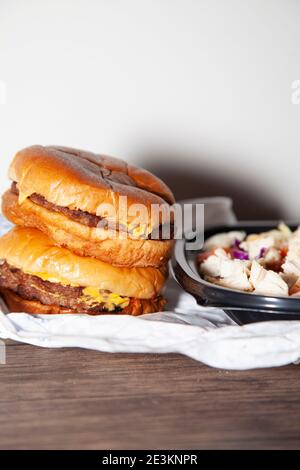 Zwei Cheeseburger gestapelt neben gegrilltem Huhn, Speck, Salat, Kohl, zerkleinerten Käse und gewürfelten Tomatensalat in einem Take-out-Behälter Stockfoto