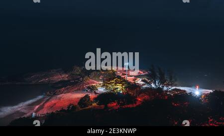 Eine Low-Key-Langzeitaufnahme mit Blick auf den Strand von Arpoador in der Nacht von oben: Küstenlinie, Flutlicht auf einem Mast, dunkler Ozeanhorizont, verschwommen t Stockfoto