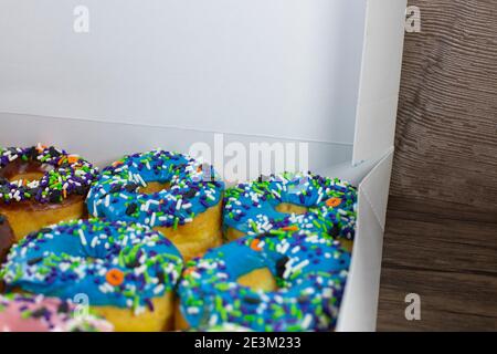 Donuts mit brauner, blauer, rosa und weißer Glasur, bedeckt mit bunten Streuseln in einer weißen Box auf einem Holztisch Stockfoto