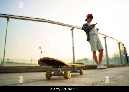 Teenage asiatische Skateboarder Junge Blick auf Handy während der Ruhe, konzentrieren Sie sich auf das Skateboard Stockfoto