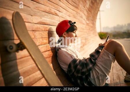 Teenage asiatische Skateboarder Junge Blick auf Handy während der Ruhe Stockfoto