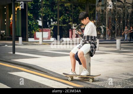 Teenage asiatische Skateboarder Kind Blick auf Handy während der Ruhe Stockfoto