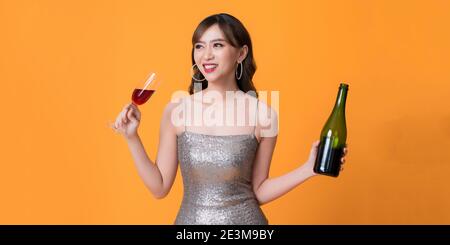 Lächelnde Frau im Abendkleid genießen Party, hält Champagner-Flasche und heben Glas auf gelbem Hintergrund Stockfoto