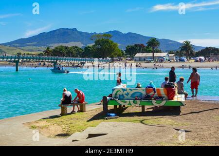 Menschen am Wasser in Raglan, einer Küstenstadt in Neuseeland, an einem heißen Sommertag. Links überquert die Te Kopua Fußgängerbrücke den Hafen Stockfoto