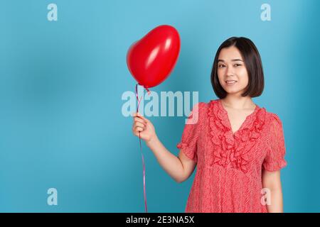 Romantische, verliebt junge asiatische Frau in rotem Kleid und dunklem Haar mit einem fliegenden roten Herz geformten Ballon isoliert auf blauem Hintergrund Stockfoto