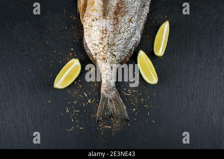 Schwanz von frischem rohem dorado auf schwarzem Schiefersteinbrett mit Gewürzen, Kräutern und Limette. Brassen Fisch bereit zum Kochen. Draufsicht. Stockfoto