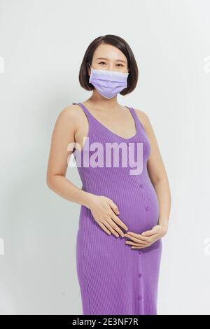 Asiatische schwangere Frau tragen Gesichtsmaske hält ihren Bauch, Blick auf Kamera, isoliert auf weißem Hintergrund, covid-19 Konzept Stockfoto