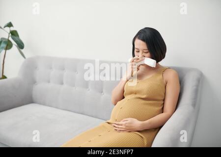 Schwangerschaft und Krankheit. Kranke schwangere Frau, die Nase in Gewebe bläst, mit Fieber, das auf dem Sofa drinnen sitzt Stockfoto