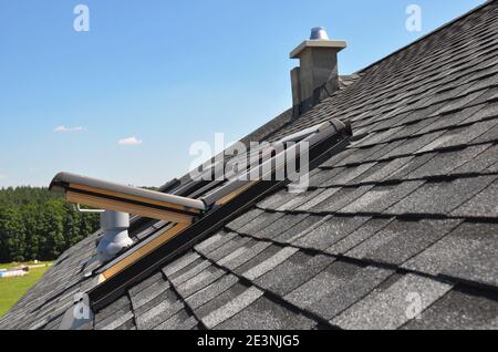 Ein graues Dach mit dimensionalen architektonischen Asphaltschindeln mit einem offenen Dachfenster, Lüftungsrohr, Luftkanal und Kamin bedeckt. Stockfoto