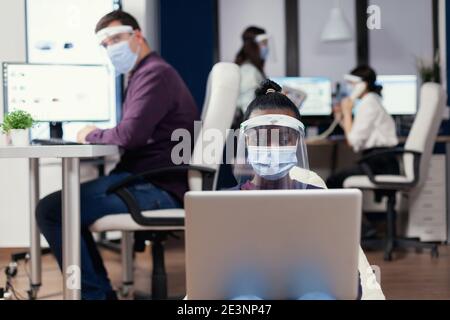 Schwarzer Manager nimmt Notizen auf Laptop trägt Gesichtsmaske gegen covid19. Multiethnisches Geschäftsteam, das während der globalen Pandemie mit Coronavirus soziale Distanz respektiert. Stockfoto