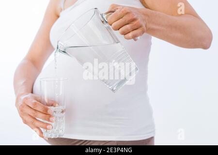 Schwangere Frau gießt Wasser aus dem Glas in das Glas Auf weißem Hintergrund Stockfoto