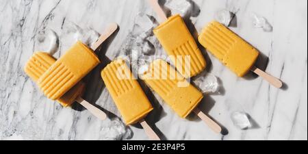 Mango Smoothie vegane Eis-Eis-Eiszapfen. Flache Mango-Fruchtpopsicles auf grauem Marmorboden unter Sonnenlicht, Draufsicht, breite Komposition. Sommer gesundes Dessert, Milchprodukte frei Food-Konzept Stockfoto