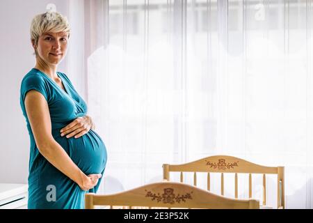 Schwangere Frau hält ihren Bauch von Kinderbett neben Das Fenster Stockfoto