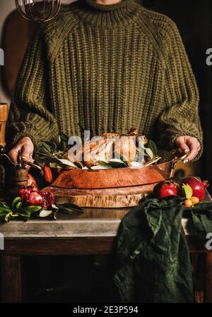 Weibchen in gestricktem Pullover hält ganze gebratenes Huhn für Winterferien festliches Abendessen in Kupfer-Bräter mit Gewürzen, Kräutern und frischem Obst. Kochkonzept Weihnachten oder Thanksgiving Stockfoto